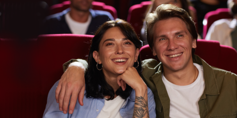 A couple smiling and laughing as they watch a performance. The man has his arm around the woman's shoulders and she's leaning into him while laughing