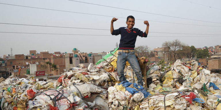 An Indian boy smiling and flexing his biceps on top of a pile of trash